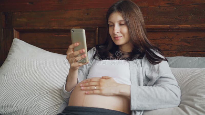 A morena grávida bonita da menina está afagando sua colisão do bebê e está usando o smartphone que relaxa na cama em casa com sor