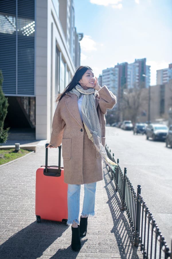 Morena hembra con abrigo beige caminando por la calle con maleta roja hablando en su móvil fotografía de archivo