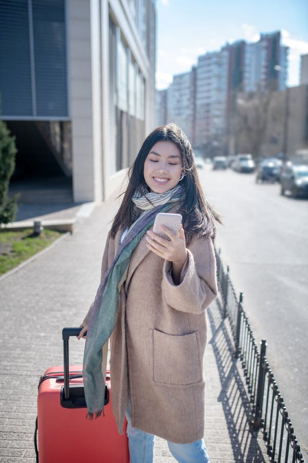 Morena hembra con abrigo beige caminando por la calle con maleta roja revisando su móvil imagen de archivo libre de regalías