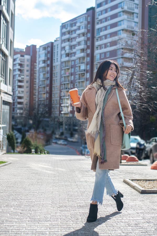 Morena hembra con abrigo beige caminando por la calle con taza de café sonriendo fotos de archivo