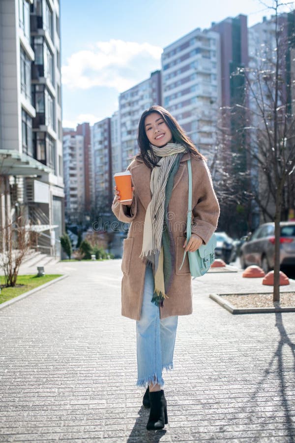 Morena hembra con abrigo beige caminando por la calle con taza de café sonriendo fotografía de archivo libre de regalías