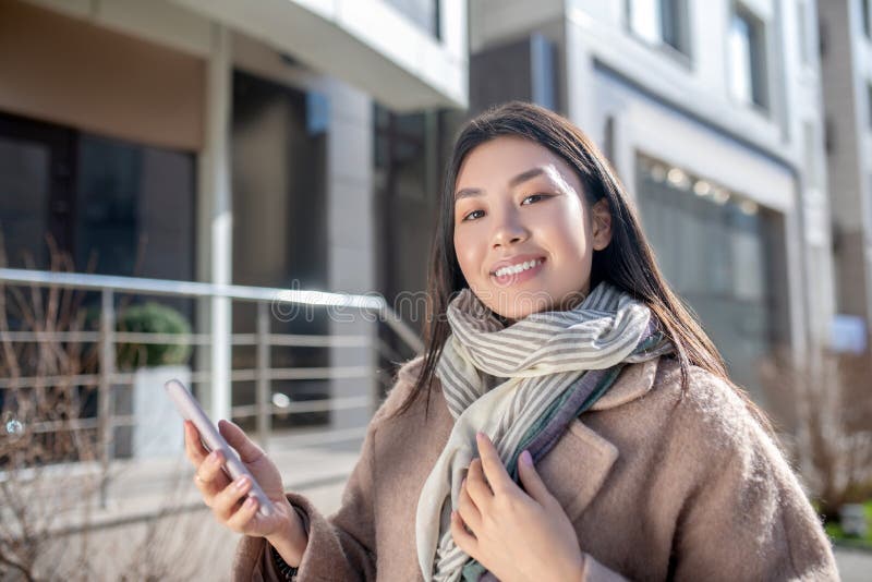 Morena hembra con abrigo beige parada afuera mirando su teléfono sonriendo fotografía de archivo libre de regalías