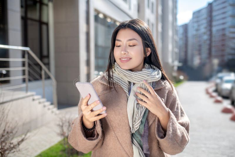 Morena hembra con abrigo beige parada afuera revisando su teléfono foto de archivo libre de regalías