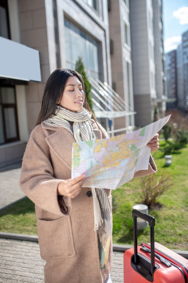 Morena hembra con abrigo beige parado en la calle con maleta roja y mapa tratando de encontrar la ubicación imagen de archivo