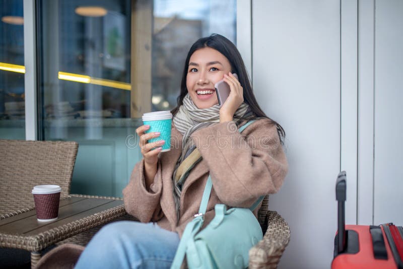 Morena hembra con abrigo beige sentada con maleta tomando café hablando por teléfono móvil imágenes de archivo libres de regalías