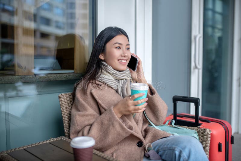 Morena hembra con abrigo beige sentada con maleta tomando café hablando por teléfono móvil fotografía de archivo libre de regalías