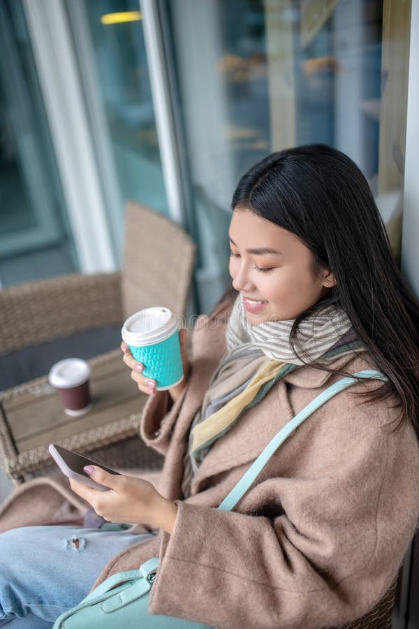 Morena hembra con abrigo beige sentada tomando café revisando su teléfono móvil imagenes de archivo