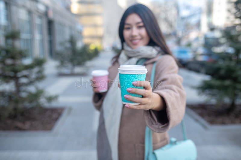 Morena hembra con abrigo beige sosteniendo tazas de café ofreciendo una taza azul a otra persona fotos de archivo