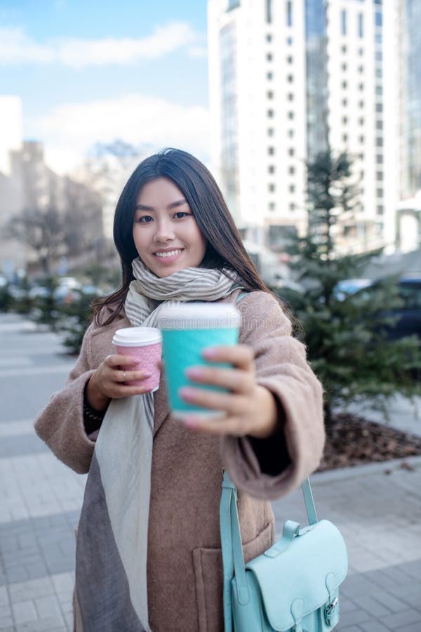 Morena hembra con abrigo beige sosteniendo tazas de café ofreciendo una taza a otra persona fotografía de archivo