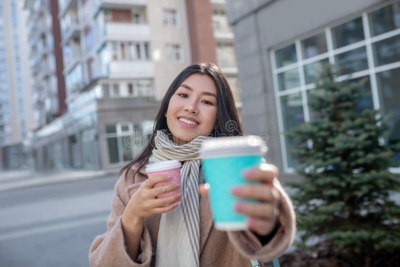 Morena hembra con abrigo beige sosteniendo tazas de café ofreciendo una taza a otra persona imagenes de archivo