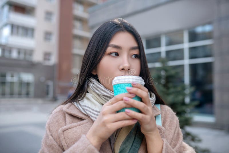 Morena hembra con abrigo beige sosteniendo una taza de café disfrutando de su bebida afuera imagenes de archivo