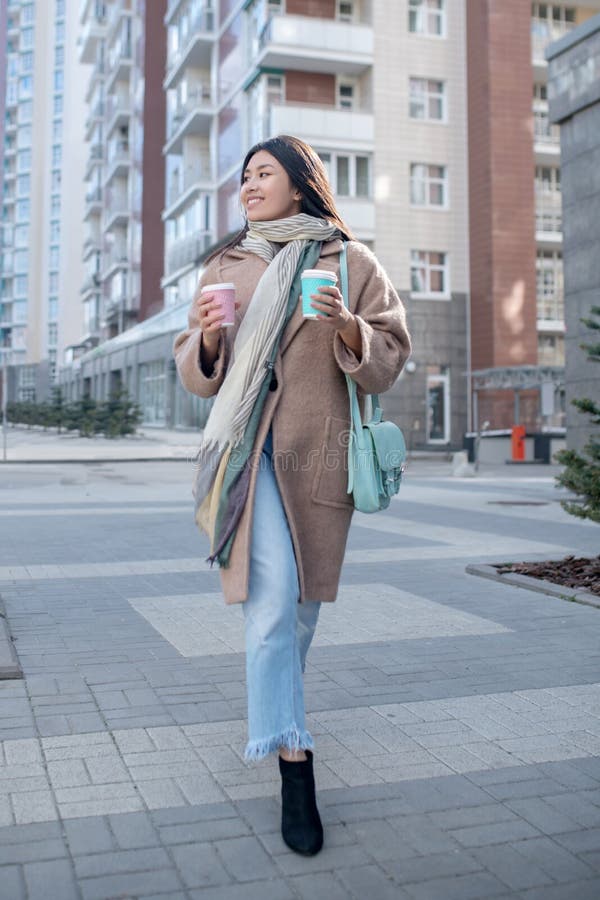 Morena hembra con abrigo beige sosteniendo una taza de café disfrutando su bebida caminando por la calle foto de archivo libre de regalías