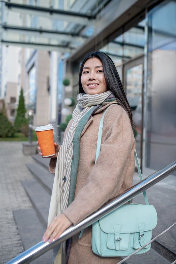 Morena hembra con abrigo beige y bolso azul sosteniendo una taza de café disfrutando de su bebida caminando por las escaleras sonr imagen de archivo libre de regalías
