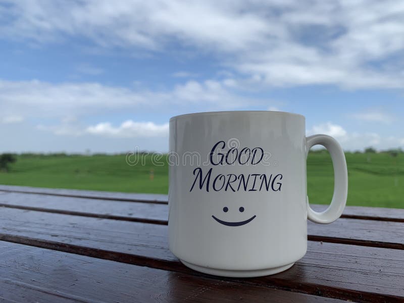 Morning greeting with a positive happy smile on white coffee or tea cup. Morning coffee concept  with white mug on the wooden table with bright summer blue sky and green field background. Good morning. Morning greeting with a positive happy smile on white coffee or tea cup. Morning coffee concept  with white mug on the wooden table with bright summer blue sky and green field background. Good morning