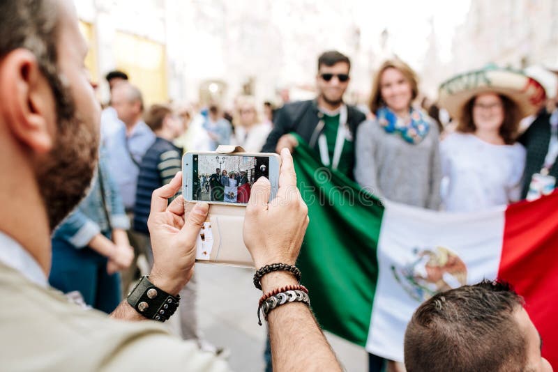 MOSKVA RYSSLAND - JUNI 2018: Händer av en man med en telefonnärbild, fotograferas han av fans av Mexico med flaggan av countren royaltyfri fotografi MOSKVA RYSSLAND - JUNI 2018: Händer av en man med en telefonnärbild, fotograferas han av fans av Mexico med flaggan av countren royaltyfri fotografi