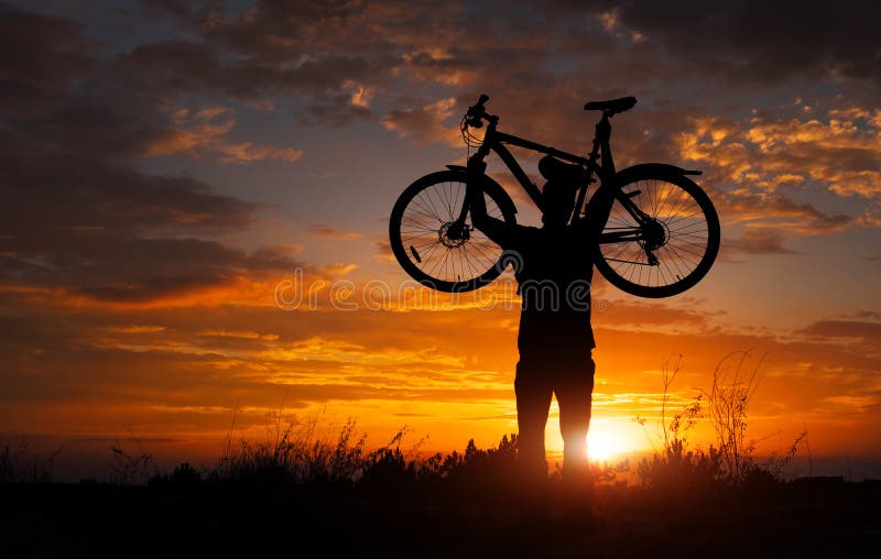 Mostre em silhueta o suporte do homem na bicicleta de levantamento da ação acima de sua cabeça imagens de stock Mostre em silhueta o suporte do homem na bicicleta de levantamento da ação acima de sua cabeça imagens de stock