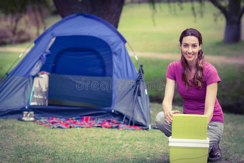 Female camper crouching on grass at campsite opening cooler beside dome tent and mug, copy space royalty free stock images
