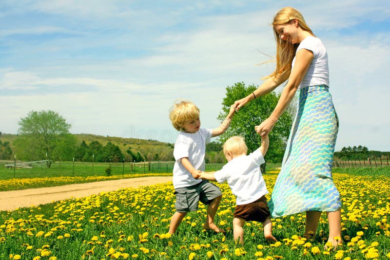 A happy mother and two children; a young boy and his baby brother, are playing outside in a meadow of Dandelion flowers, holding hands and dancing on a Spring day. A happy mother and two children; a young boy and his baby brother, are playing outside in a meadow of Dandelion flowers, holding hands and dancing on a Spring day.