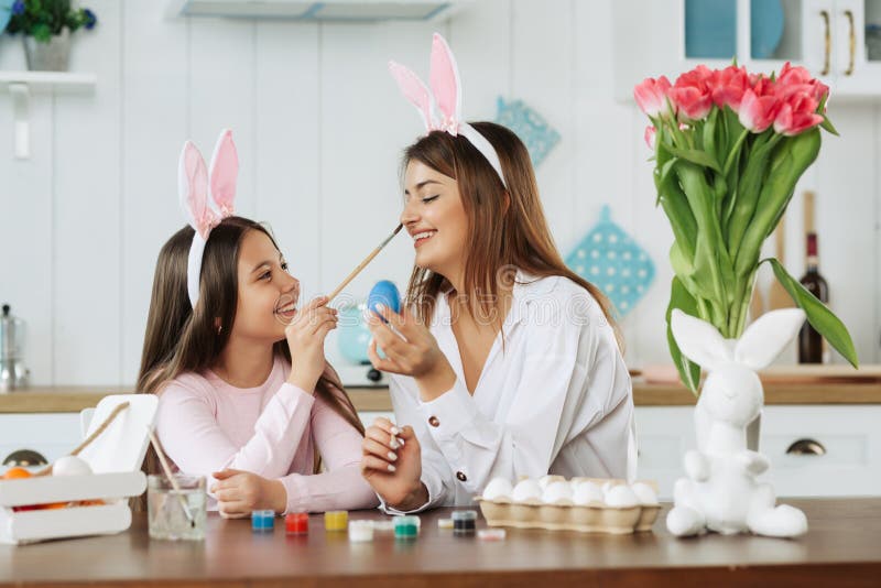 A mother and daughter celebrating Easter, painting eggs with brush. Happy family smiling and laughing, drawing on face. Cute little girl in bunny ears preparing the holiday
