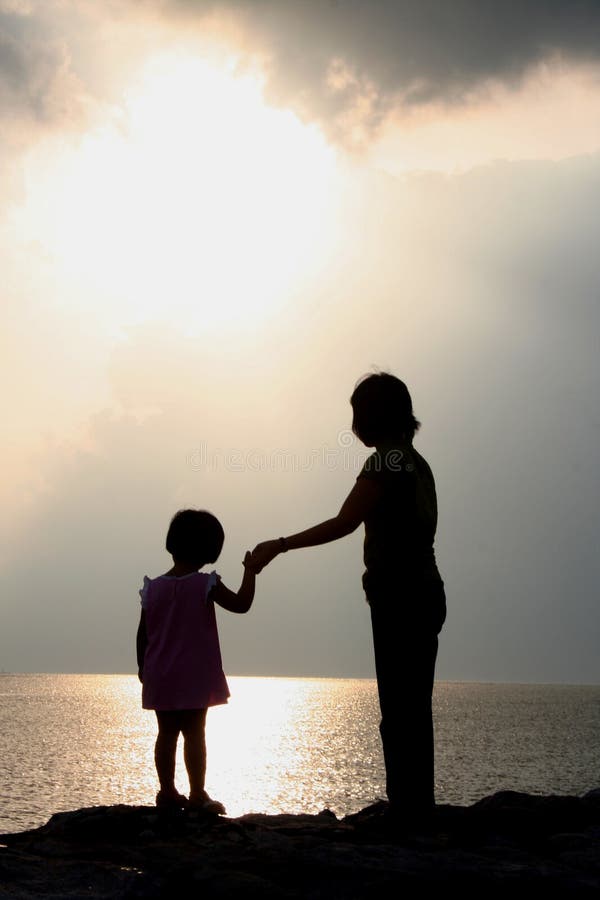 A mother and daughter stand silhouetted, holding hands as they watch the sunset from a sandy beach overlooking the ocean. A mother and daughter stand silhouetted, holding hands as they watch the sunset from a sandy beach overlooking the ocean.