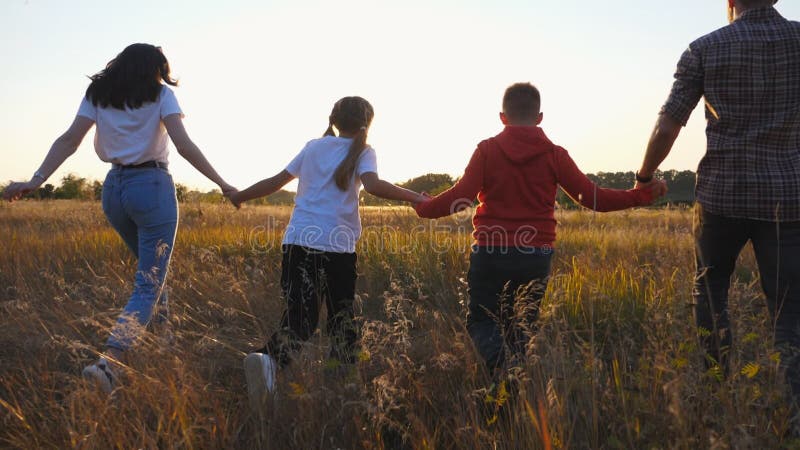 Mother and father with two kids holding hands of each other and running through grass field at sunset. Happy parents