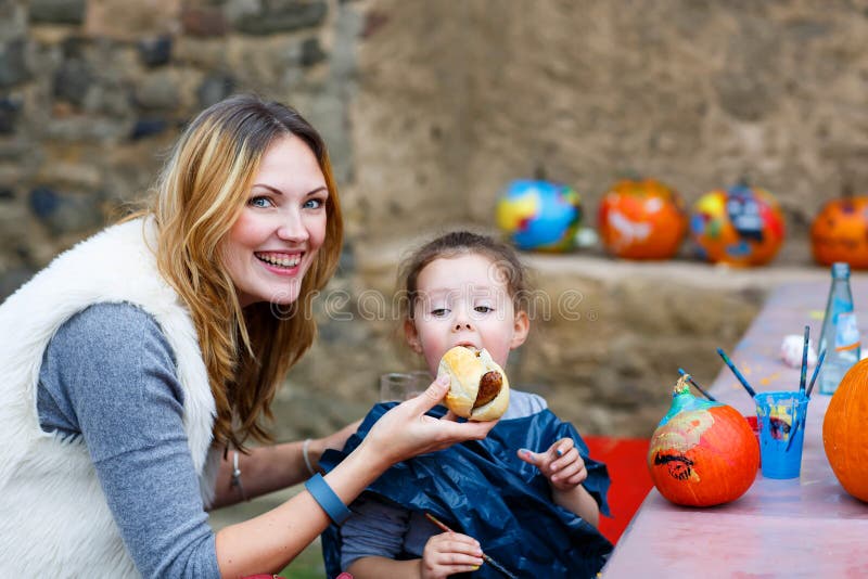 Young beautiful mother feeding her little daughter, adorable kid girl with hot dog or German sausage on thanksgiving or halloween pumpkin festival, outdoors. Family enjoy warm autumn day. Young beautiful mother feeding her little daughter, adorable kid girl with hot dog or German sausage on thanksgiving or halloween pumpkin festival, outdoors. Family enjoy warm autumn day.