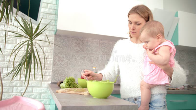 Mother holding baby on hands in kitchen. Mom with kid prepare dinner