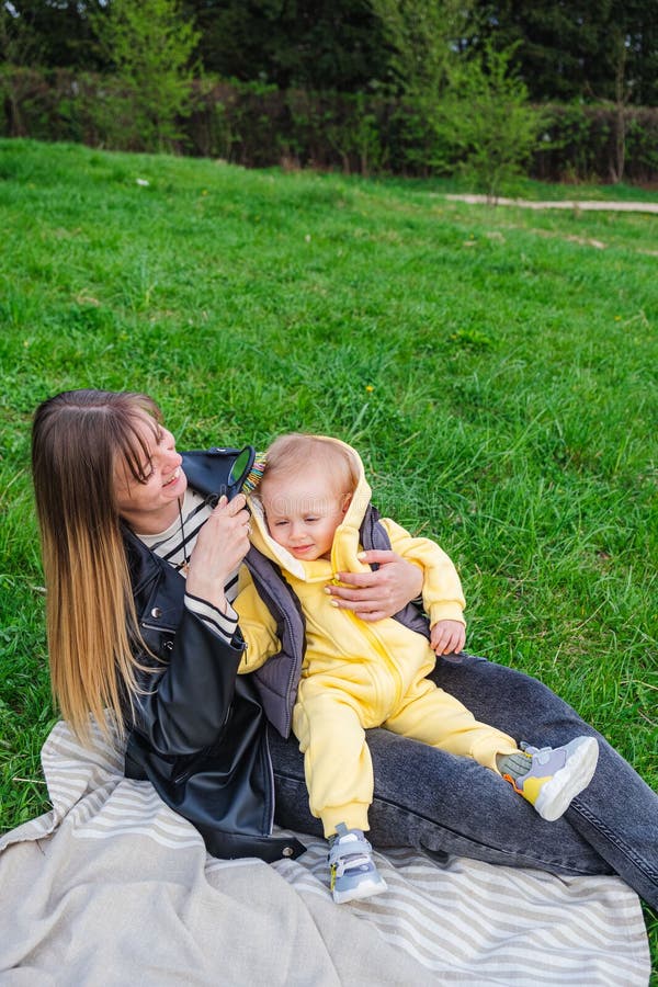 Mama își ține fiul în poală și stă pe o pătură pe un gazon verde într-un parc de primăvară stock photography