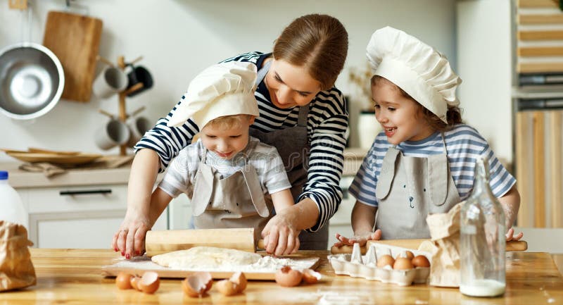 Happy young mother and little kids in chef hats rolling dough together while preparing cookies in home kitchen. Happy young mother and little kids in chef hats rolling dough together while preparing cookies in home kitchen