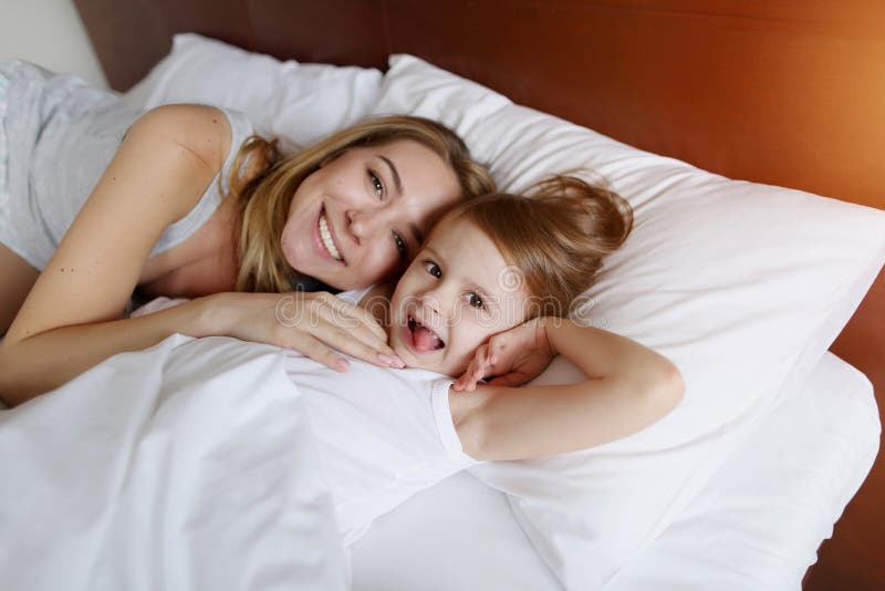 Mother and little daughter look at camera smiling on white bed with sunshine