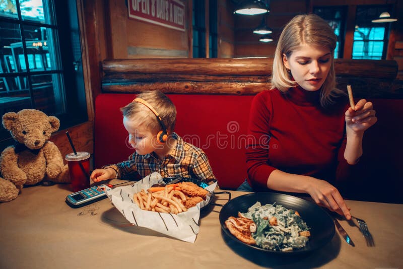 Mother and toddler boy son at restaurant. Caucasian family mother with toddler boy son eating food dinner in cafe restaurant and playing electronic digital royalty free stock image