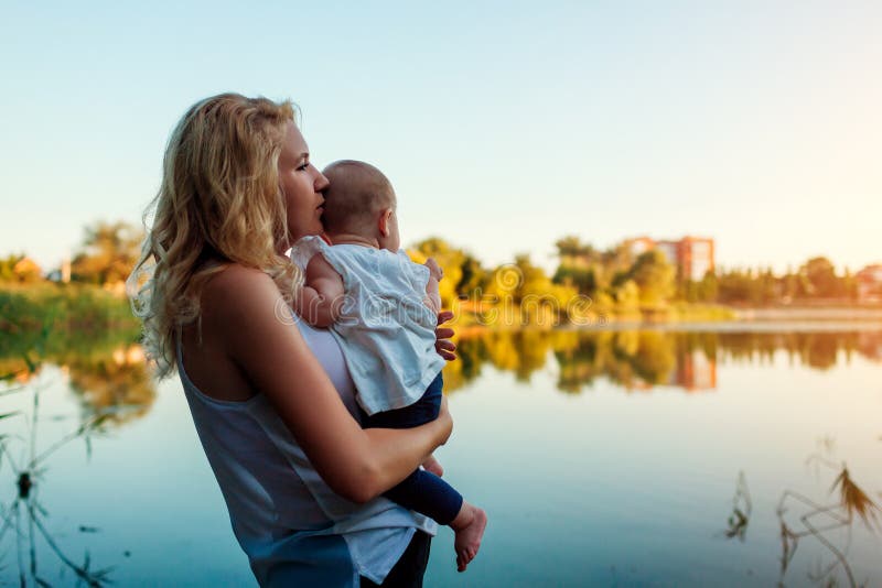 Mothers day. Young woman holding and hugging baby girl by spring river. Family walking outdoors. Kid exploring world