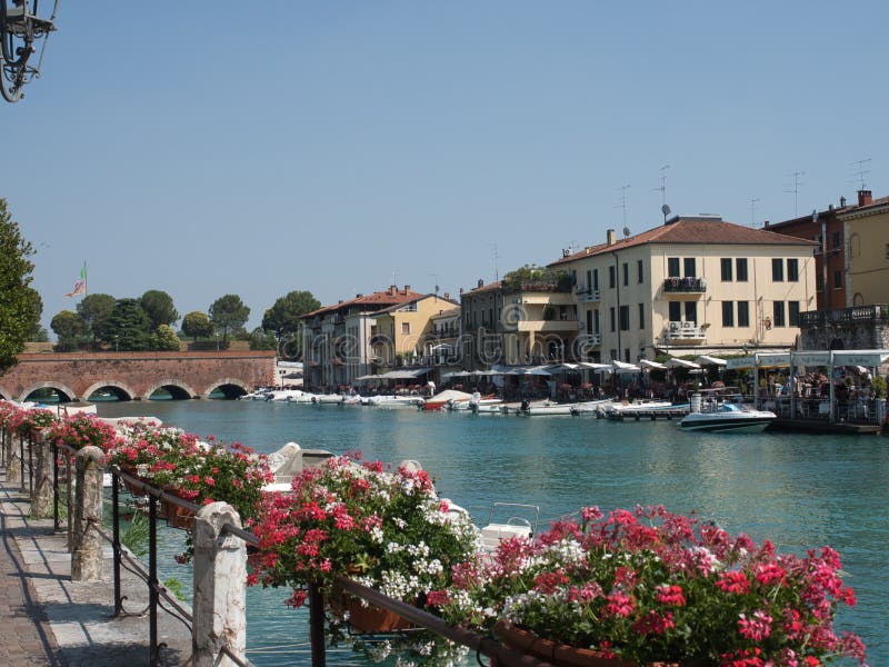 Motorbåtar som parkeras längs den peschieradel garda kanalen i sjögardaen, Italien med lokala affärer i bakgrunden och blommorna fotografering för bildbyråer Motorbåtar som parkeras längs den peschieradel garda kanalen i sjögardaen, Italien med lokala affärer i bakgrunden och blommorna fotografering för bildbyråer