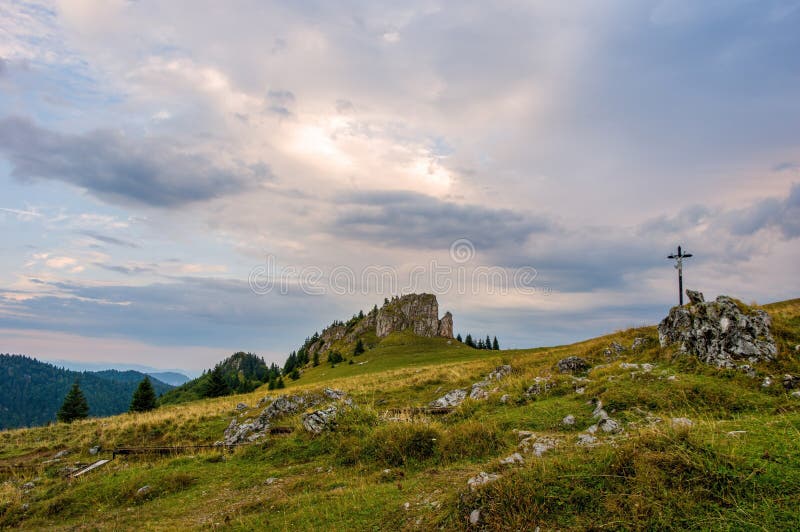 Horská krajina se skalami, pastvinami pro dobytek a malým křížkem v pozadí.