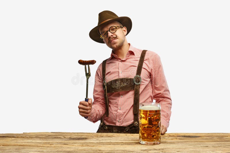 Bavarian mustache man in hat wearing traditional fest outfit holding hot fried sausage and looking at huge glass of beer. Concept of Oktoberfest, traditions, drinks and food. Copy space for ad. Bavarian mustache man in hat wearing traditional fest outfit holding hot fried sausage and looking at huge glass of beer. Concept of Oktoberfest, traditions, drinks and food. Copy space for ad