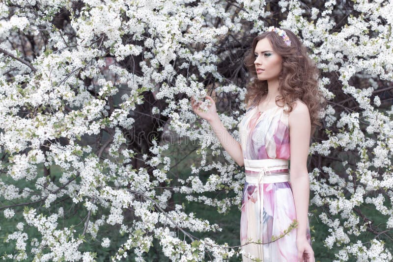 Moça bonita com ondas em um vestido colorido ar com uma grinalda em sua cabeça que anda no parque perto de uma flor de cerejeira imagens de stock Moça bonita com ondas em um vestido colorido ar com uma grinalda em sua cabeça que anda no parque perto de uma flor de cerejeira imagens de stock