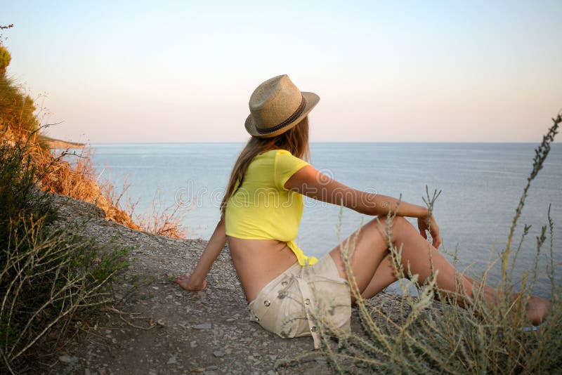A moça está sentando-se em uma costa de mar rochoso selvagem e está olhando-se o mar no por do sol fotos de stock A moça está sentando-se em uma costa de mar rochoso selvagem e está olhando-se o mar no por do sol fotos de stock