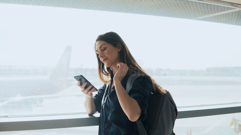 Moça que usa o smartphone perto da janela do aeroporto A mulher europeia feliz com trouxa usa o app móvel no terminal 4K