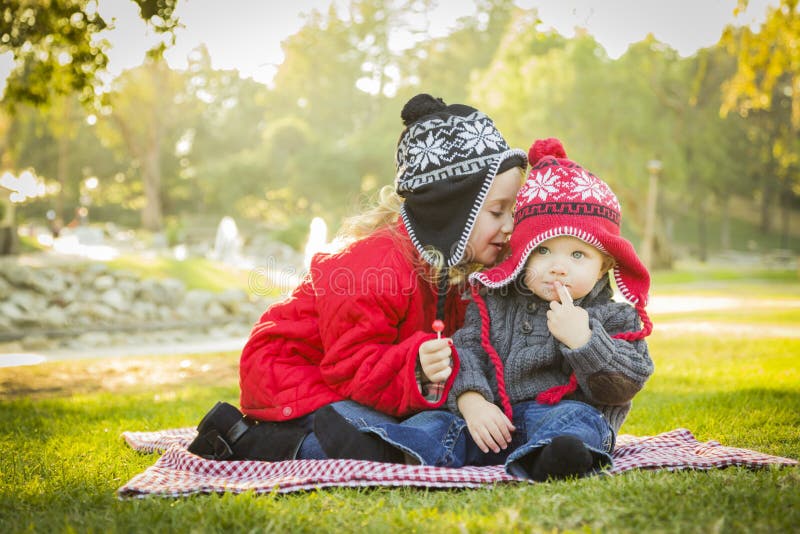 Little Girl Whispers A Secret to Her Baby Brother Wearing Winter Coats and Hats Sitting Outdoors at the Park. Little Girl Whispers A Secret to Her Baby Brother Wearing Winter Coats and Hats Sitting Outdoors at the Park.