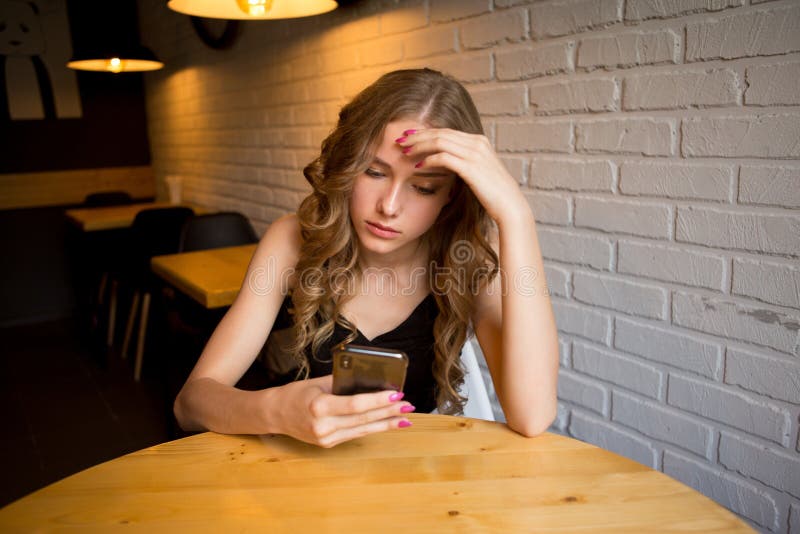 Moça triste que senta-se em um café que parece cansado seu telefone, menina infeliz que olha seu telefone preto