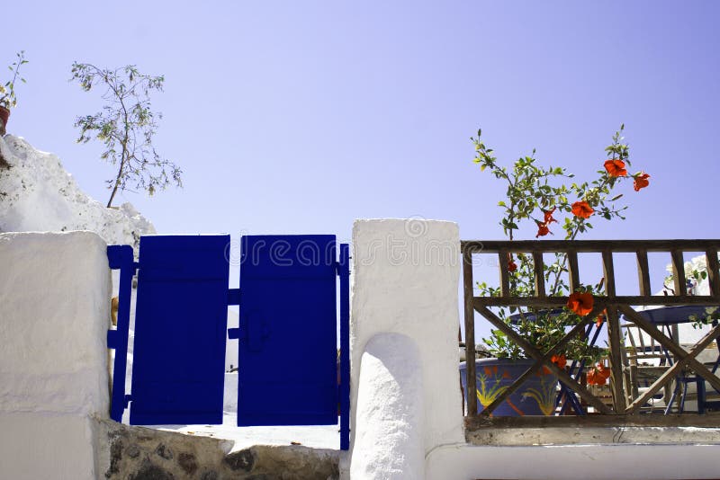 Blue wooden painted gate and white garden wall with blooming flowers and sky background, Santorini Island, Greece. Blue wooden painted gate and white garden wall with blooming flowers and sky background, Santorini Island, Greece.