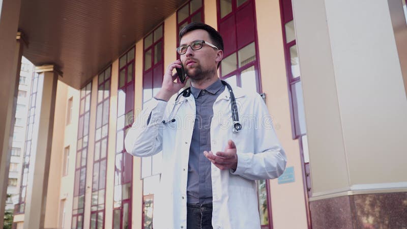 MS physician in white lab coat speaking by cell phone outside clinic. Healthcare worker outdoors talking on mobile phone