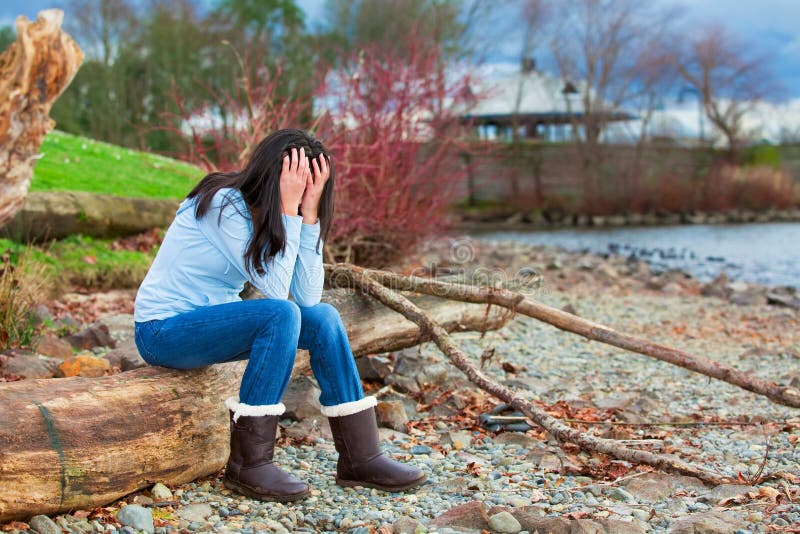 Muchacha adolescente joven triste que se sienta en registro a lo largo de la playa rocosa por el lago imágenes de archivo libres de regalías Muchacha adolescente joven triste que se sienta en registro a lo largo de la playa rocosa por el lago imágenes de archivo libres de regalías