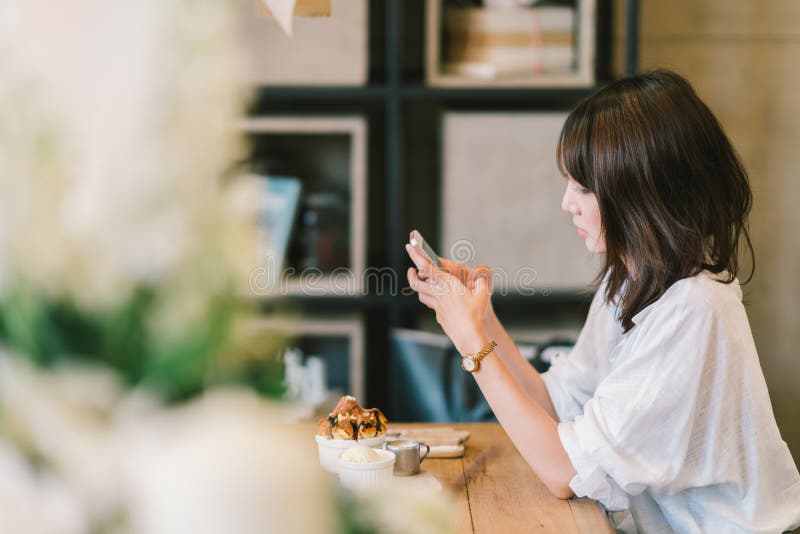 Muchacha asiática hermosa que usa smartphone en el café con la tostada del chocolate y el helado Postre de la cafetería y forma d imagenes de archivo