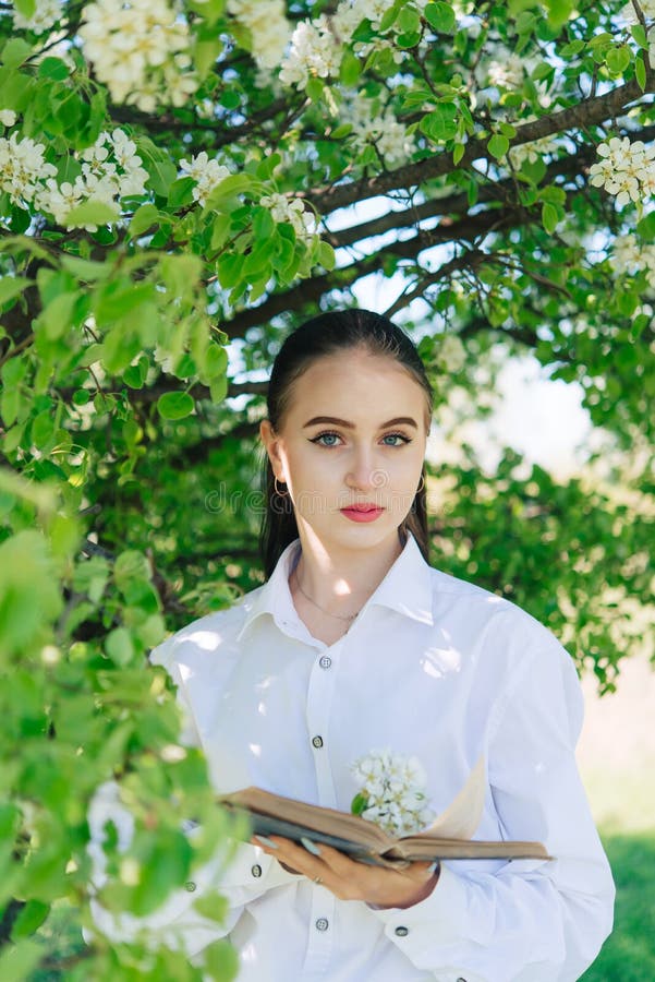 Girl in a white blouse and blue jeans. Stands near a flowering tree and reads a book. Girl in a white blouse and blue jeans. Stands near a flowering tree and reads a book.