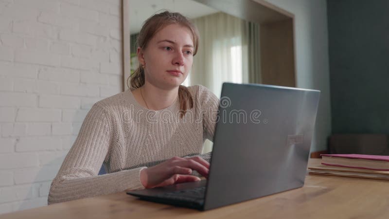 Muchacha con un acogedor suéter trabajando en un ordenador portátil en casa sentada en una mesa de madera con libros de texto en u almacen de metraje de vídeo