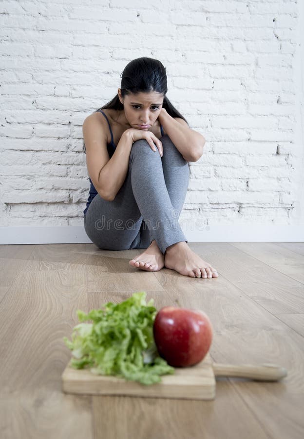 Young woman or teenager girl sitting on ground alone and worried at home suffering nutrition and eating disorder in diet and weight loss obsession with healthy food in anorexia and bulimia concept. Young woman or teenager girl sitting on ground alone and worried at home suffering nutrition and eating disorder in diet and weight loss obsession with healthy food in anorexia and bulimia concept