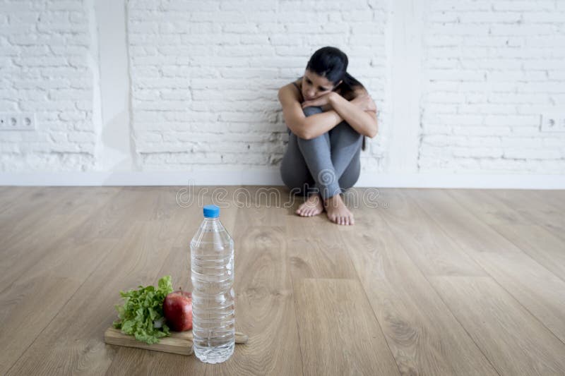 Young woman or teenager girl sitting on ground alone and worried at home suffering nutrition and eating disorder in diet and weight loss obsession with healthy food in anorexia and bulimia concept. Young woman or teenager girl sitting on ground alone and worried at home suffering nutrition and eating disorder in diet and weight loss obsession with healthy food in anorexia and bulimia concept