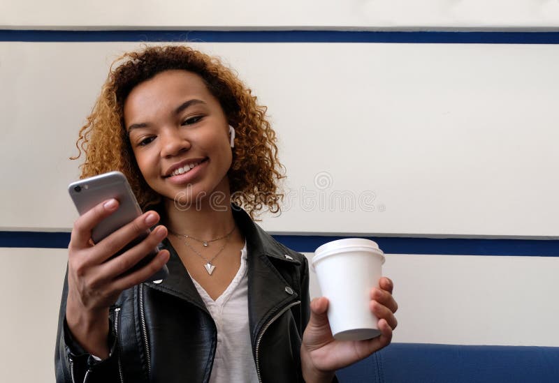 Muchacha de piel morena hermosa feliz que sostiene un vidrio blanco en su mano y que mira en el teléfono Un auricular inalámbrico fotografía de archivo Muchacha de piel morena hermosa feliz que sostiene un vidrio blanco en su mano y que mira en el teléfono Un auricular inalámbrico fotografía de archivo