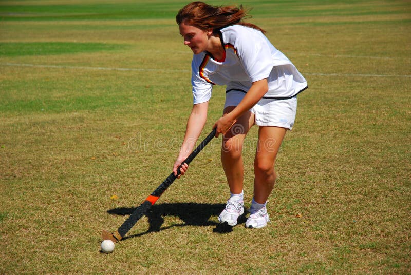 A beautiful active caucasian white Hockey player girl with concentrating facial expression hitting the ball on the field outdoors. A beautiful active caucasian white Hockey player girl with concentrating facial expression hitting the ball on the field outdoors